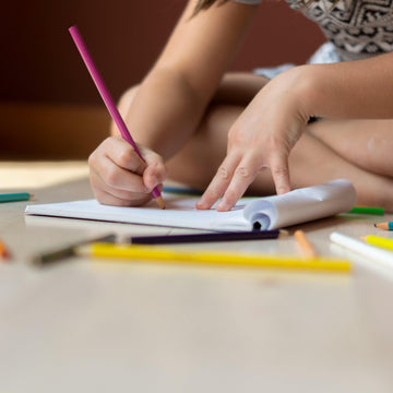 Child writing with a pink pencil on a notebook surrounded by colorful pencils on a wooden surface.